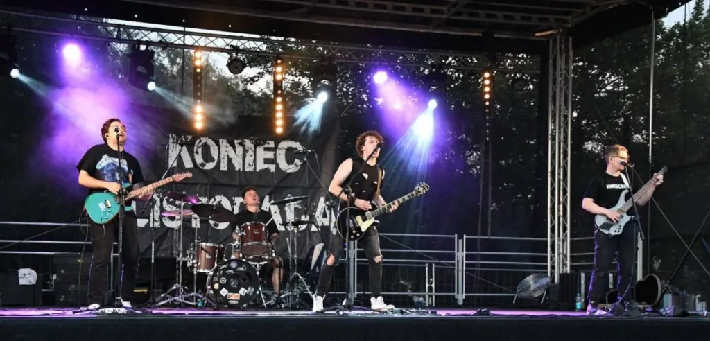 Zespół rockowy występujący na scenie plenerowej na placu w centrum miasta, tłum ludzi przed sceną, gitary, perkusja, letni wieczór