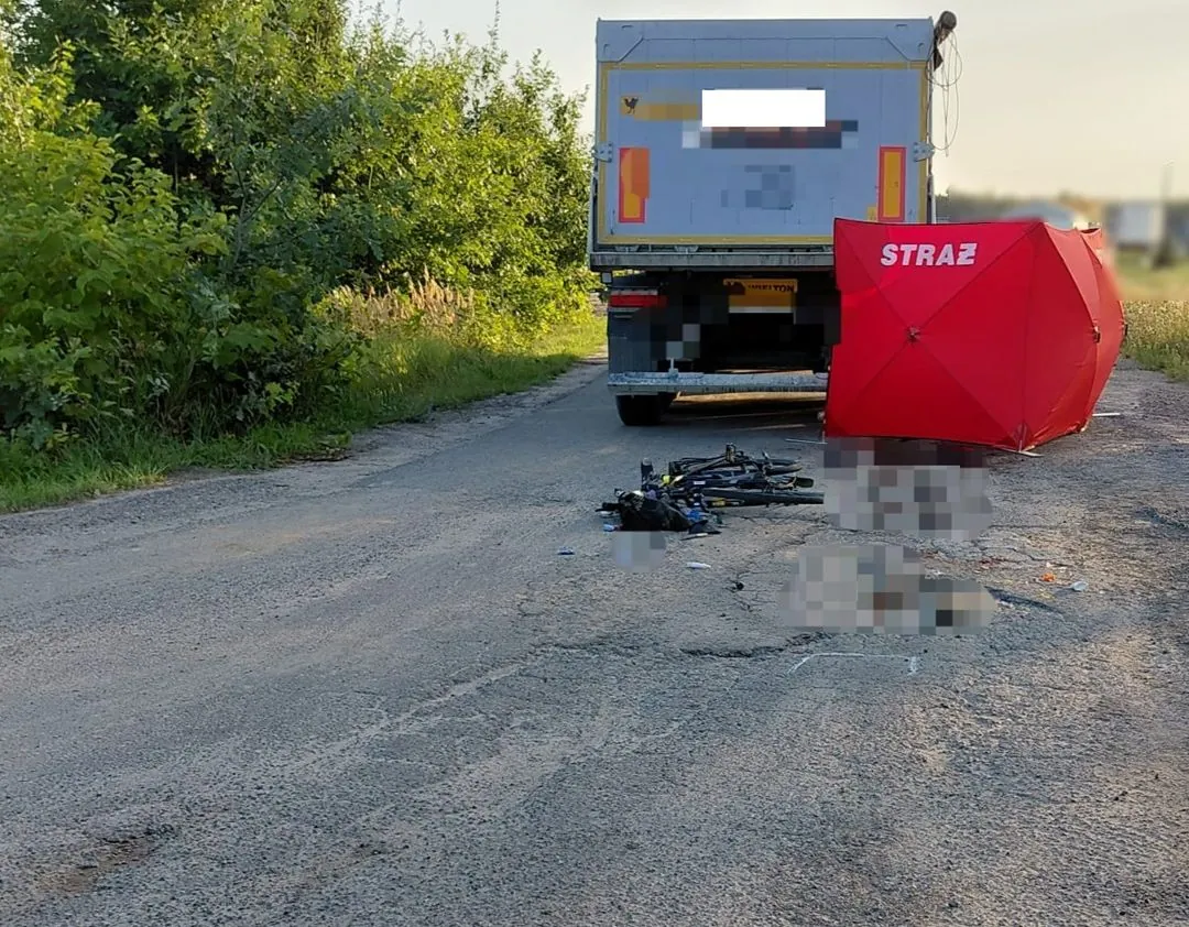 Miejsce wypadku z udziałem ciężarówki i rowerzystki, policja i karetka na miejscu, rower leżący na drodze, ciąg pieszo-rowerowy.