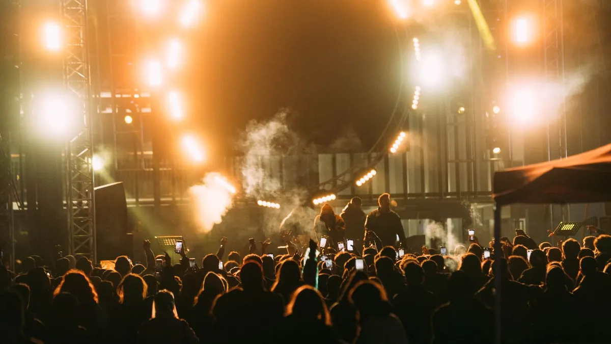 Tłum ludzi stoi przed sceną oświetloną jasnymi reflektorami i światłami w ciepłych barwach.