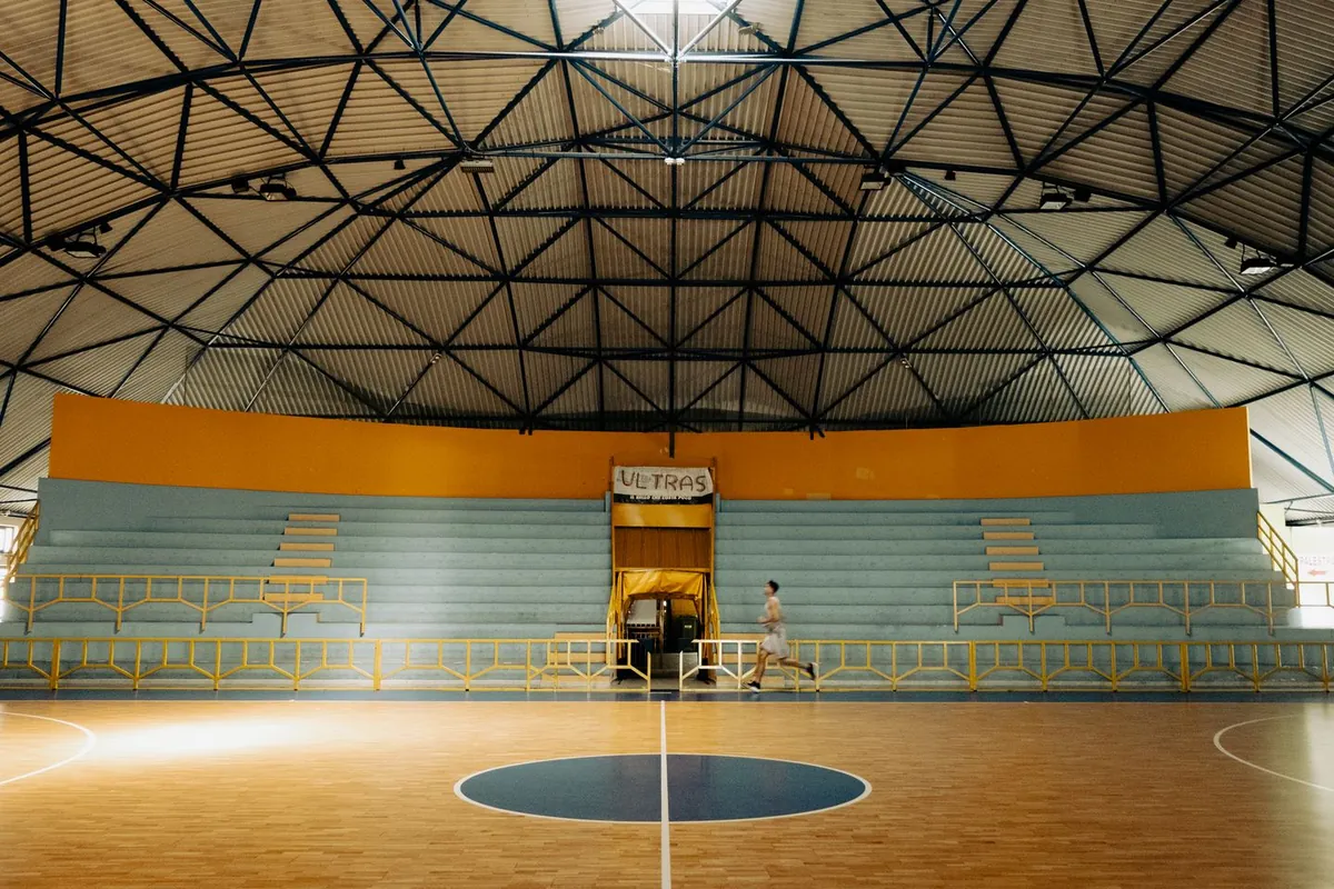 Wnętrze hali sportowej z drewnianym boiskiem, niebieskimi trybunami i dwiema małymi osobami przy środku.