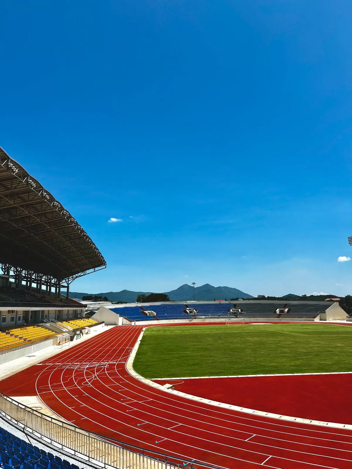 Pusty stadion z czerwoną bieżnią wokół zielonej murawy, trybunami i górami na horyzoncie.
