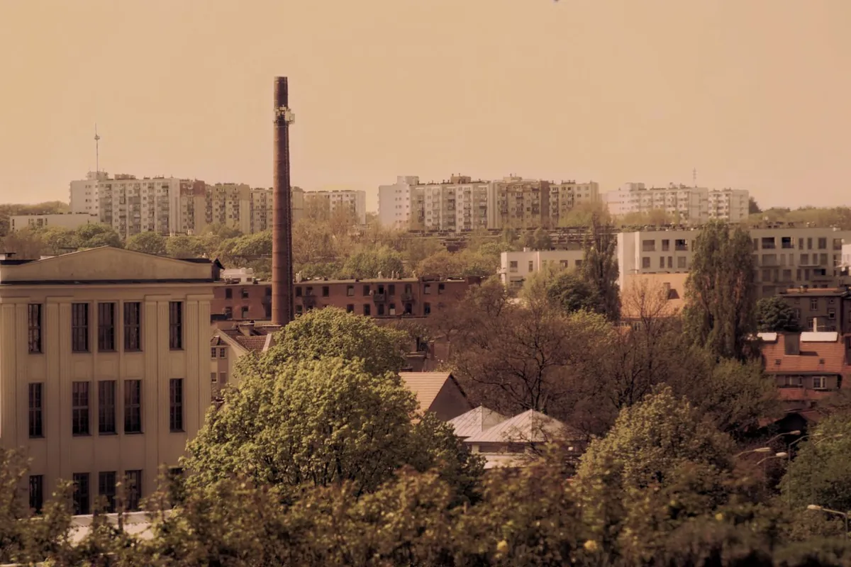 Panorama zabudowy miejskiej z blokami mieszkalnymi w tle, kominami i drzewami na pierwszym planie.