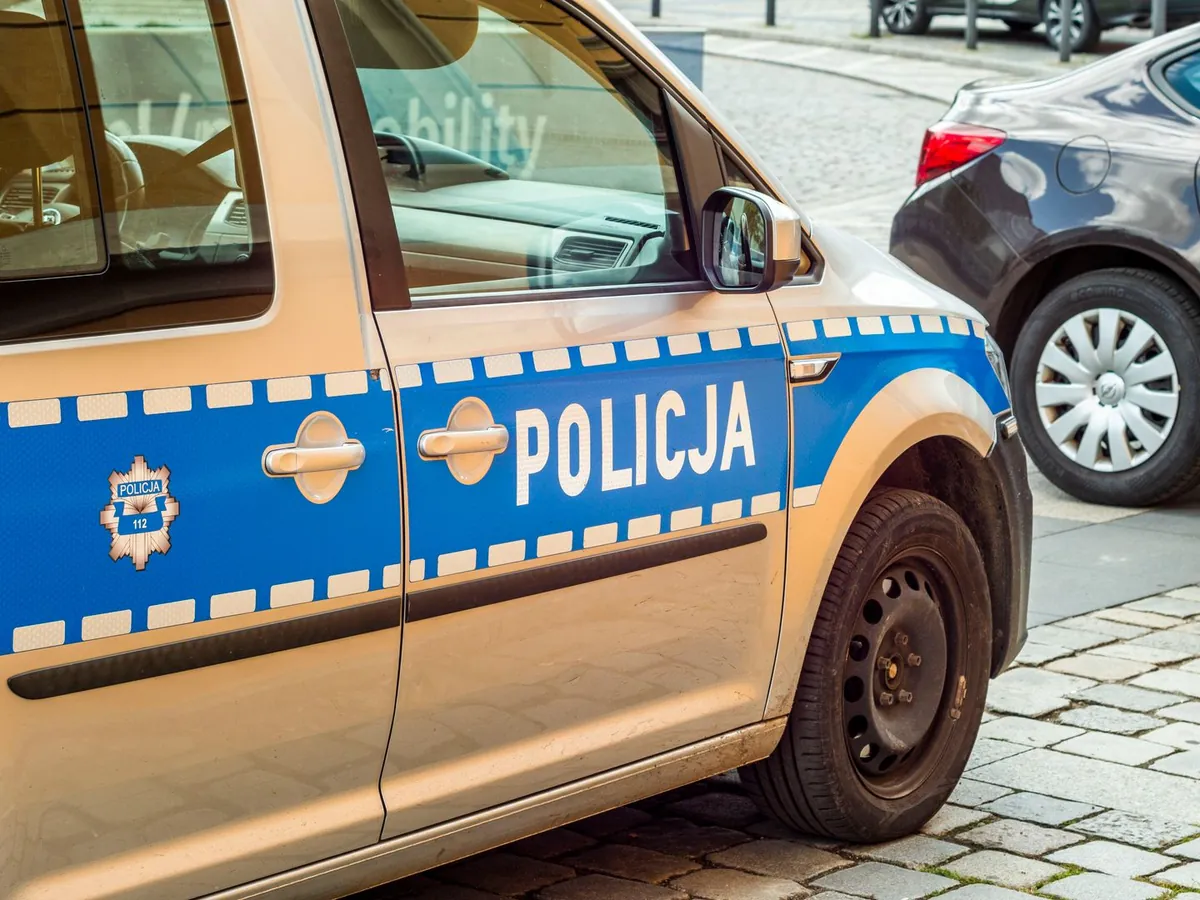 Zbliżenie białoniebieskiego samochodu policyjnego z napisem „POLICJA”, zaparkowanego przy ulicy obok innego samochodu.