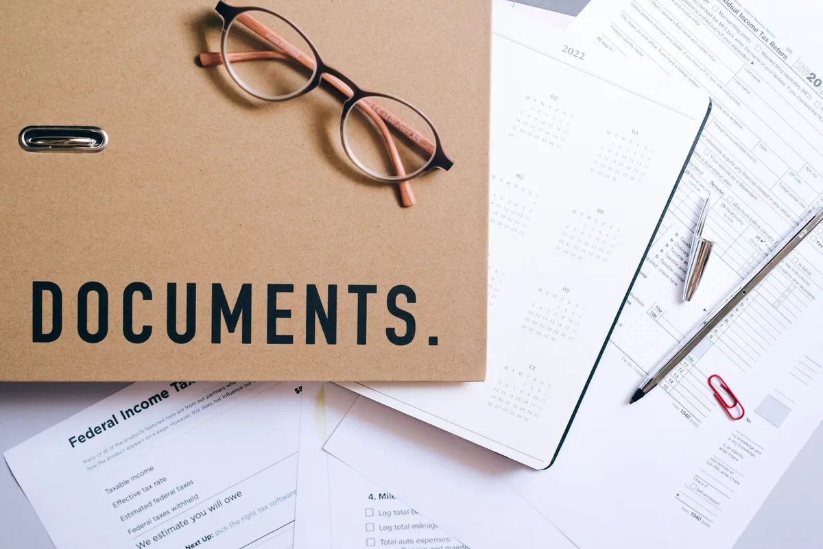 Na dokumentach i formularzach leżą okulary, długopisy oraz teczka z napisem „DOCUMENTS”.