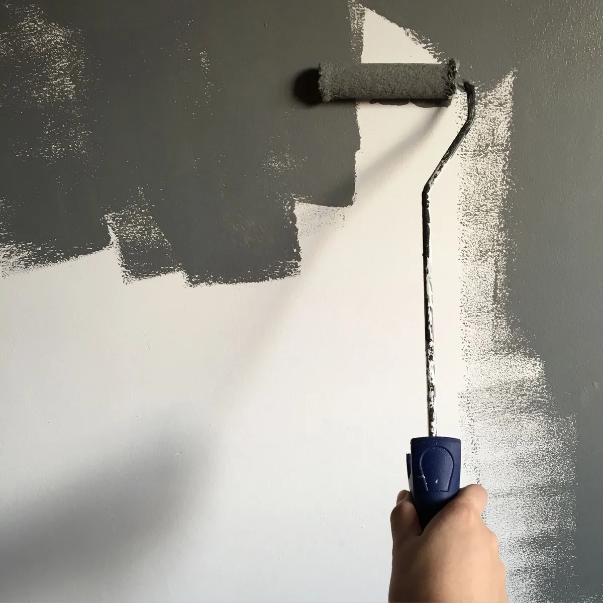 Dłoń prowadzi wałek z szarą farbą po białej ścianie.